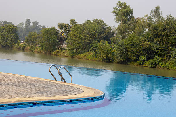 Luxury resort infinity swimming pool featuring a curved edge and tiled deck, offering a panoramic view of the natural body of water and surrounding lush tropical greenery. Perfect vacation setting.