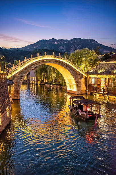 Chinese water town glowing with warm lights under a twilight sky, showcasing traditional architecture, a historic stone bridge, and a tour boat navigating the calm canal