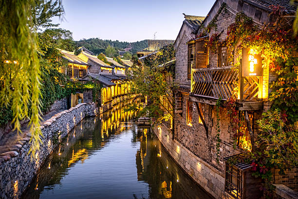 Traditional Chinese buildings with intricate architecture and vibrant autumn foliage are illuminated by warm lights, reflecting in the calm waters of a canal through a historic water town