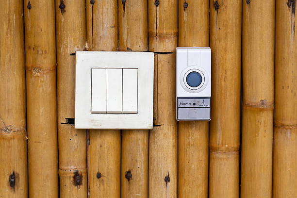Close-up of a white 3-gang electrical light switch and modern doorbell unit installed side-by-side on a rustic, woven bamboo wall texture. Contrast of new technology and traditional materials.