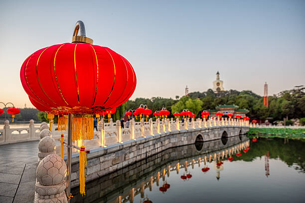 Red lanterns are lighting a classic stone bridge over a calm lake, reflecting light and structures beautifully, with the iconic White Dagoba dominating the background during dusk
