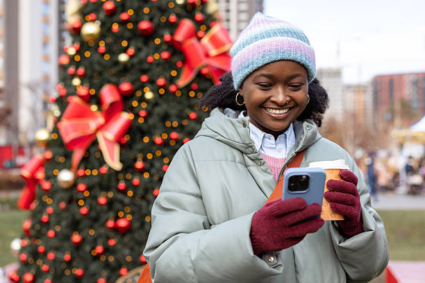 istock Young woman smiling and using smartphone outdoors by decorated Christmas tree 2251211071