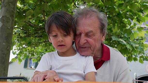 Elderly man and young boy focused on reading a book together outdoors, sharing a tender and educational moment under the shade of trees. intergenerational bonding