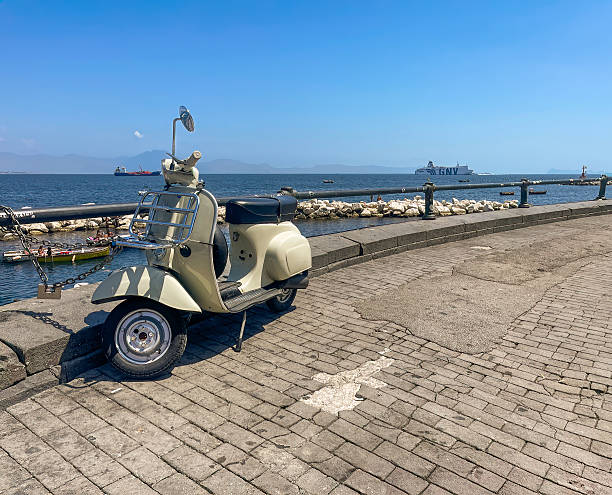 Naples, Italy - July 17, 2024: Via Nazario Sauro Rotonda. Bathers on the rocks and a classic Vespa scooter parked. GNV ferry boat on the sea