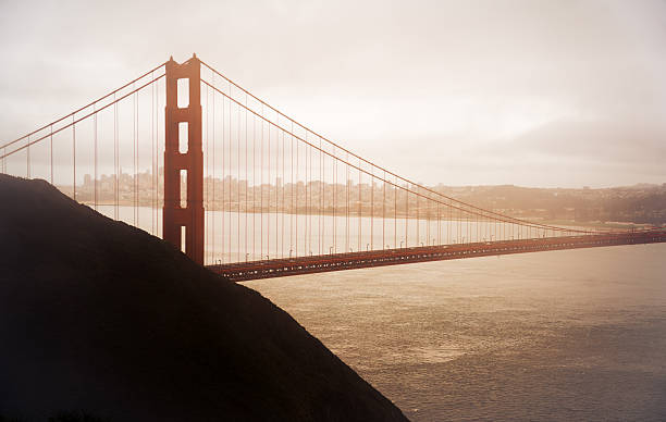 A stunning warm-toned photograph of the Golden Gate Bridge rising through soft atmospheric haze at sunrise. The iconic red suspension bridge stretches across San Francisco Bay, with the city skyline visible in the background under dramatic glowing light. Shot from a high vantage point, this landscape highlights the bridge’s architectural beauty, the peaceful water, and the golden California ambiance. Perfect for themes related to travel, tourism, landmarks, engineering, transportation, and West Coast scenery