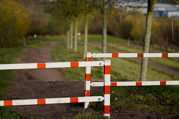 Closeup barrier across tree lined path sunlit park avenue with red and white post, shallow depth of field isolates painted metal,