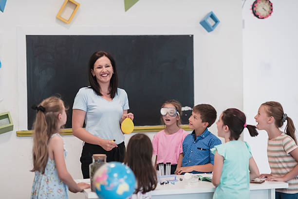 istock Elementary School Science Classroom: Enthusiastic Teacher Explains Chemistry to Diverse Group of Children, Little Boy Mixes Chemicals in Beakers. Children Learn with Interest 2251230176