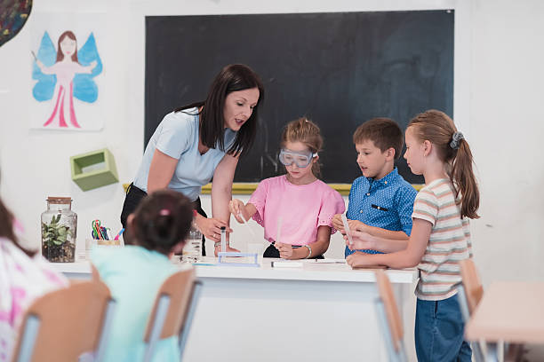 istock Elementary School Science Classroom: Enthusiastic Teacher Explains Chemistry to Diverse Group of Children, Little Boy Mixes Chemicals in Beakers. Children Learn with Interest 2251232193