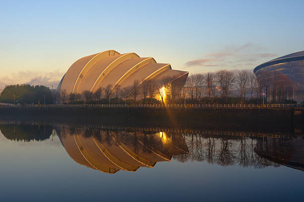 The iconic SEC Armadillo and OVO Hydro Arena are beautifully reflected in the calm waters of the River Clyde in Glasgow, Scotland. Captured during golden hour, the modern architecture stands out against the soft light and cloudy sky, creating a striking urban landscape. This image showcases a prominent landmark and event venue within the vibrant cityscape.