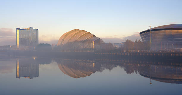 A serene view of the SEC Armadillo and the Hydro Arena reflected in the calm waters of the River Clyde in Glasgow, Scotland. The modern architecture stands out against the soft morning light, creating a picturesque urban landscape. The scene evokes a sense of tranquility and showcases Glasgow's iconic landmarks.