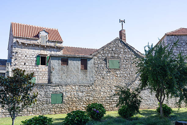 A historic stone building with green shutters, surrounded by trees and a manicured garden. Bright daylight highlights textured walls, roof tiles, and a tranquil, rustic countryside vibe.