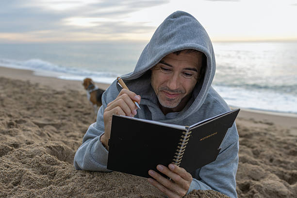 Man in a grey hoodie lying on sandy beach, writing in a black notebook and reflecting, finding peace and inspiration by the ocean with his beagle dog nearby