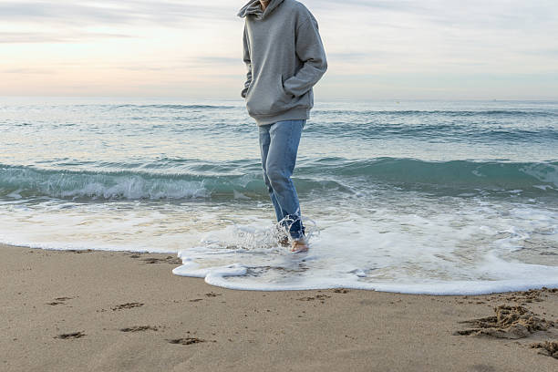 Individual wearing a grey hoodie and jeans stepping into tranquil ocean waves at the shore, splashing water while strolling on wet sand with bare feet during daytime