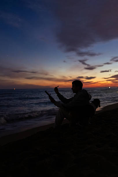 man sitting on the beach at sunset, silhouetted against a vibrant sky, quietly journaling and sketching while watching the ocean waves and horizon