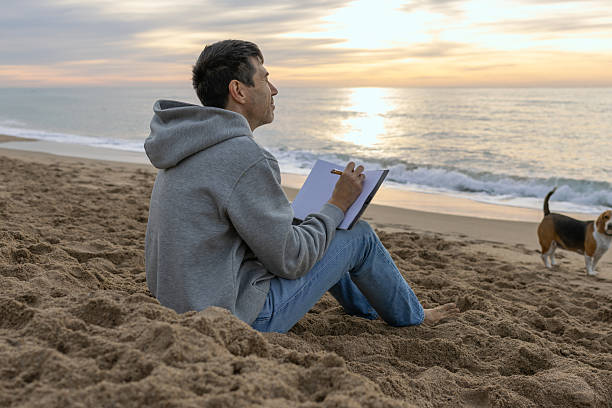 Mid adult man journaling and reflecting on sandy beach with his beagle dog during a beautiful sunrise or sunset, finding inspiration and peace by the ocean