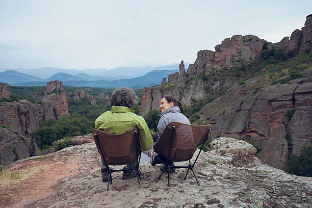 Two senior hikers are sitting on outdoor chairs and enjoying the view of the valley in Belogradchik Rocks, Bulgaria. Explorers couple on adventure travel