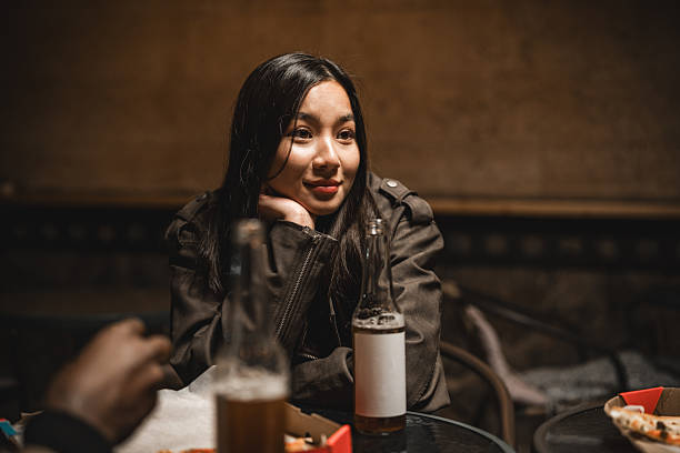 In a cozy atmosphere, a young woman rests her chin on her hand, deep in thought while surrounded by friends and drinks. The soft lighting creates an intimate vibe perfect for conversation.