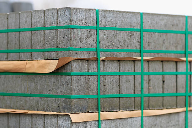 Heavy concrete blocks are stacked neatly, secured with green straps and brown paper, awaiting transportation for a construction project. The setting is a construction yard.