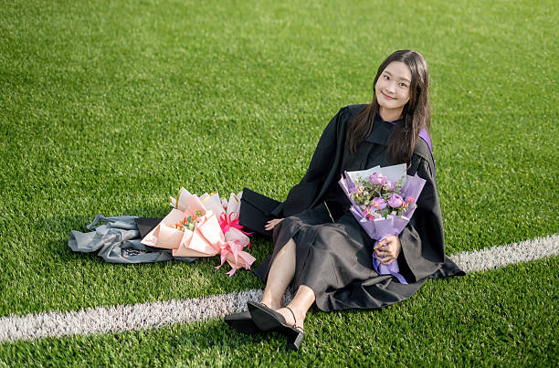 A graduate student in a robe sits on the grass, holding bouquet and enjoying a joyful moment after the ceremony