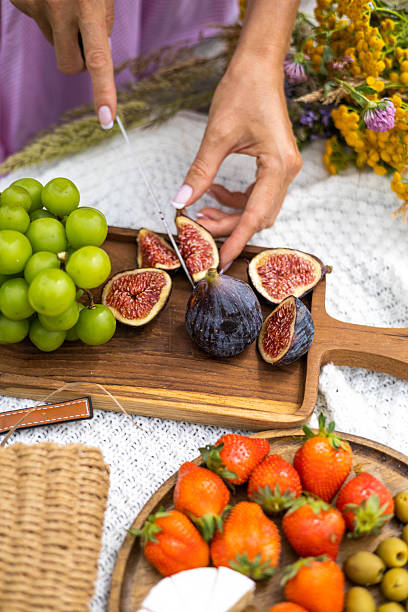 Hands slicing fresh figs on wooden board during summer picnic with grapes, strawberries, olives