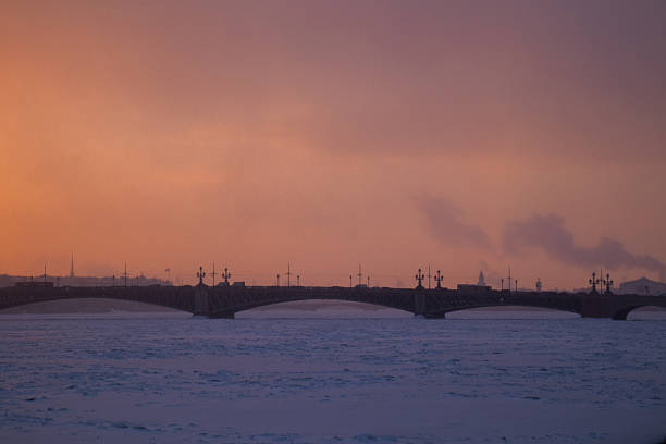 Silhouette of a historic bridge over the frozen Neva River and the Saint Petersburg skyline under a wide, dramatic orange and pink winter sunset sky