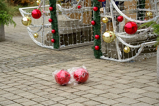 Two large red Christmas tree balls in plastic packaging lying on the paving stones in front of a decorative Christmas structure.