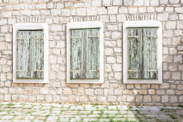 A rustic stone wall features three green wooden shutters, worn and peeling. The faded boards add charm to the exterior, with a cobblestone path and sparse grass hinting at a quiet, aged building.