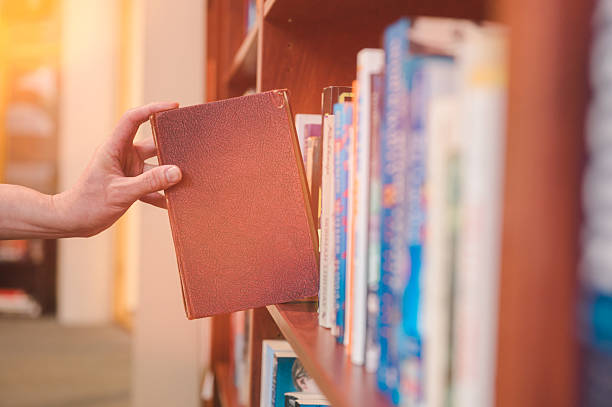 istock Close-up of a person’s hand selecting a red book from a library shelf with sunlight streaming through the window, symbolizing education, learning, knowledge, and reading inspiration 2251298537