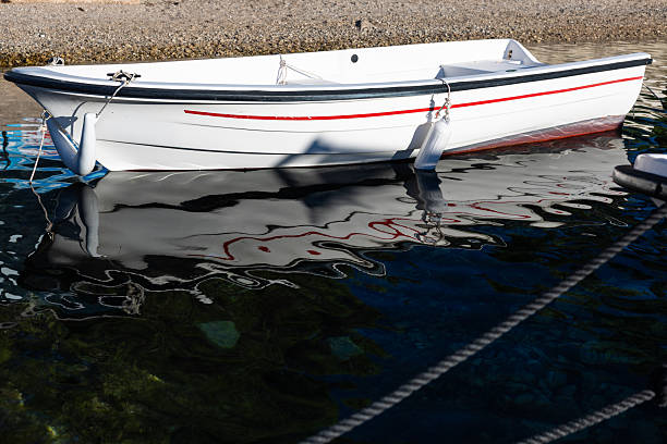 A white boat rests at the shoreline, its red stripe mirrored in the calm water. Ropes and fenders hang at the hull, creating a tranquil, coastal scene perfect for travel and leisure themes.