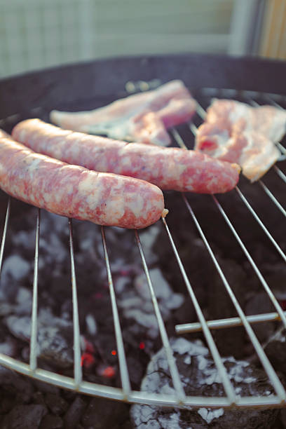 Sausages and two bacon slices cooking on a charcoal barbecue close up