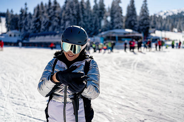 A man points his hand at the camera in sunglasses with a ski helmet on his head. and holds a stick in his hand against the background of white snow and mountains..