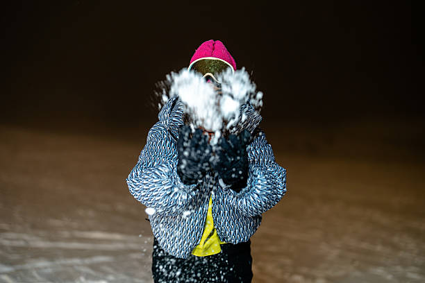 A person enthusiastically interacts with snow, captured during a nighttime winter scene. Wearing a stylish jacket and hat, their movement conveys a sense of joy and energy, embodying the charm of snowy evenings. The setting is serene with a blurred snowy background.