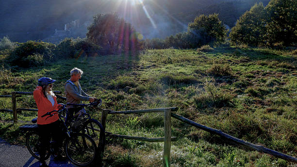 Mature couple with e-bikes pause by agricultural field and fence, Liguria