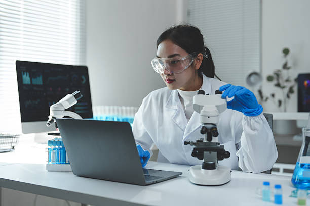 istock Asian female scientist wearing lab coat and gloves, using laptop and adjusting microscope while conducting research in modern laboratory with test tubes and computer displaying data analysis 2251307530