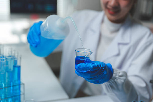 istock Scientist wearing lab coat and gloves pouring blue liquid reagent from plastic bottle into beaker glass during experiment in chemical laboratory 2251307626