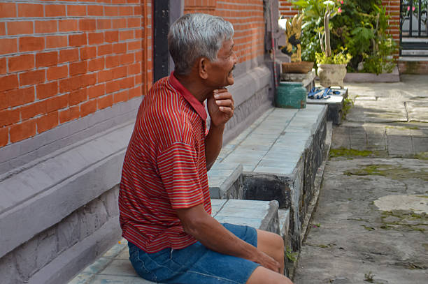An elderly man in a red and white striped polo shirt and blue shorts sits on a raised tiled terrace next to a brick wall, resting and gazing off to the side with his hand on his chin.