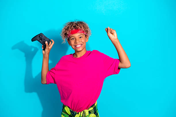 A happy young boy with curly hair and a red bandana holds a game controller in a cheerful pose against a blue backdrop, conveying fun playfulness and casual lifestyle.