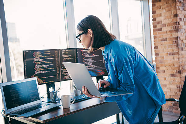 istock Businesswoman programmer works on laptop beside multiple monitors in a modern loft office with brick wall 2251317017