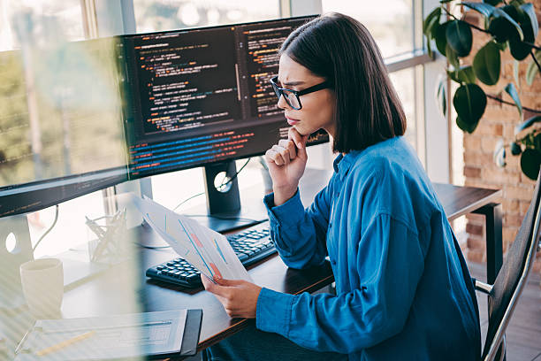 istock Female programmer reviews charts and code at a bright loft office with dual monitors and papers in hand 2251317909