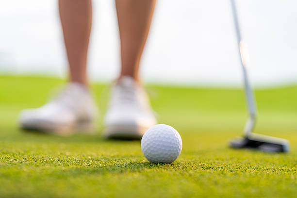Close up of a woman stands on a well-maintained golf green, concentrating as she prepares to putt. The putter and golf ball are clearly visible, highlighting her focus and dedication to the game.