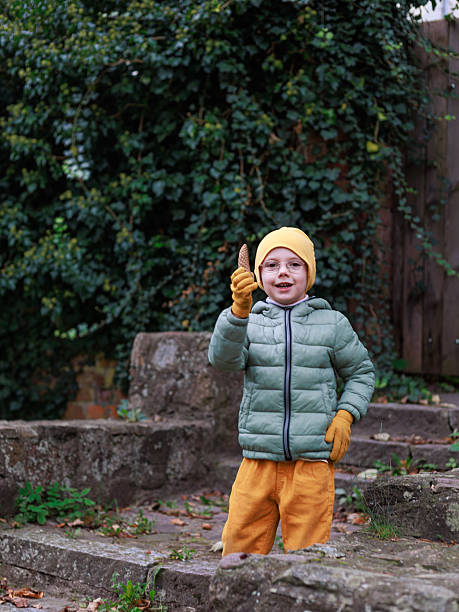 istock A smiling child in bright autumn clothes holds up a pine cone while standing on old stone steps against a textured green ivy background with open copy space. Concept of playful childhood, nature discovery, autumn mood and outdoor joy. 2251323793