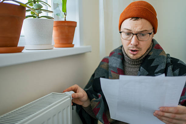 istock Man feeling cold and anxious, wearing winter clothes indoors while holding a large utility bill with a surprised expression, concerned about rising energy expenses 2251327476