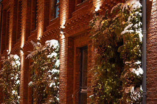 Brick building exterior showing evergreen trees covered in fresh snow and adorned with glowing christmas lights, highlighting a festive and cozy celebration during a cold winter night