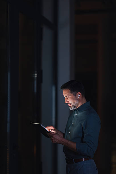 istock Evening Office Worker Checks Tablet With Wireless Earbuds in Dim Hallway 2251332301
