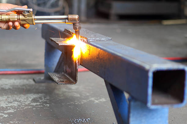 A hand holds a brass cutting torch, generating intense flame and bright sparks while precisely heating and cutting a heavy steel I-beam in a dark workshop.