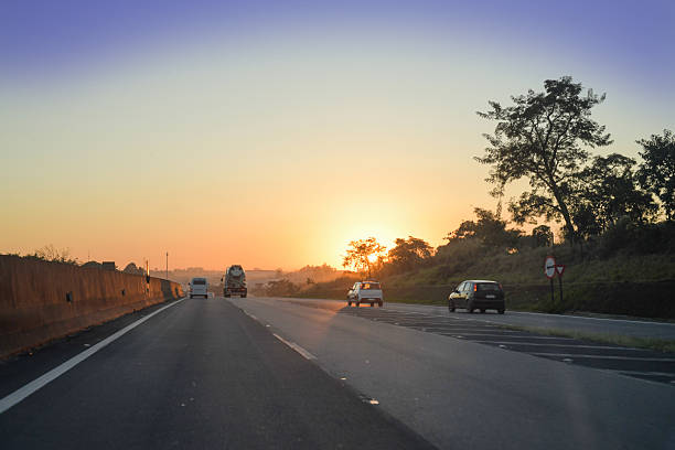 Road scene at sunrise, with the sun emerging behind a tree-covered hill, casting soft light across the landscape. The natural illumination, tree silhouettes, and peaceful atmosphere convey a sense of travel, freedom, and the beauty of the early morning.