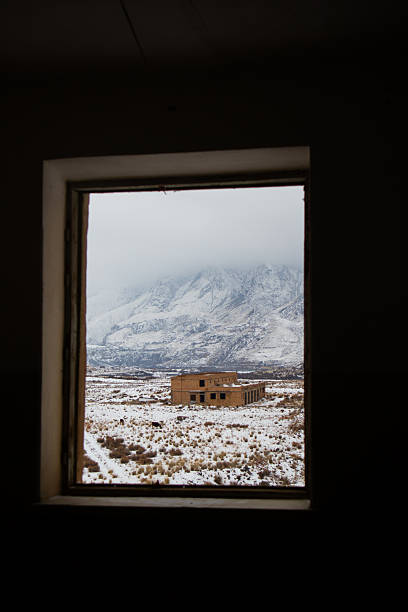 Looking through a rectangular window frame at a ruined brick building in a snowy valley with mountains in the background