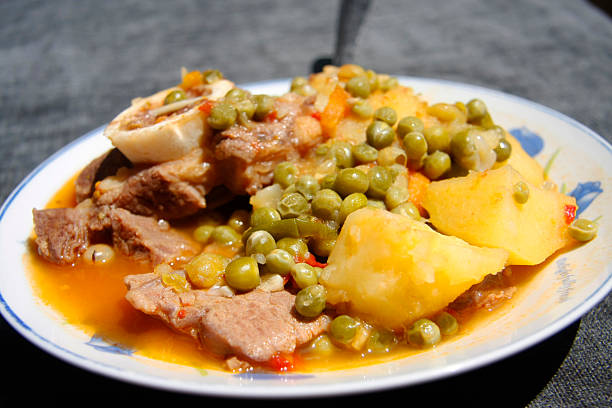 A close-up, high-angle shot of a white plate containing a traditional stew, likely Carne con Papas y Guisantes (Meat with Potatoes and Peas). The dish features chunks of cooked meat, yellow potato pieces, and green peas in a savory, orange-colored broth. Ideal for themes of global cuisine, comfort food, recipes, traditional cooking, and Latin American or Spanish food blogs/cookbooks.