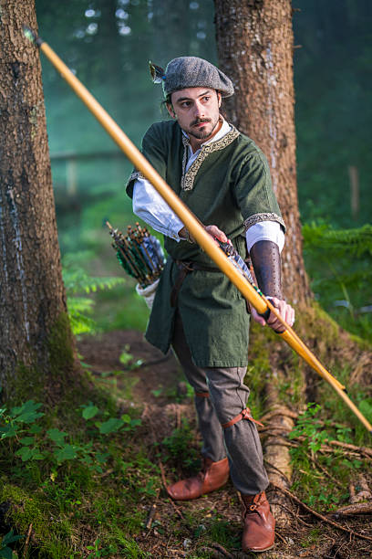 Young Caucasian male archer in medieval dress stands in a forest, holding a bow with intent focus. The lush greenery and surrounding trees create a serene backdrop as he prepares for the next move with concentration and poise.