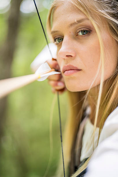 Young Caucasian woman in medieval dress standing in a wooded area, aiming her bow with a look of concentration as she prepares to release the arrow.